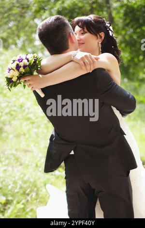 Felice coppia di sposi che abbraccia la natura, regge un bouquet, sorride Foto Stock