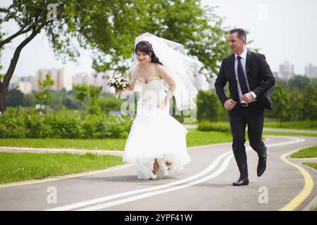 La sposa e lo sposo corrono insieme lungo una strada asfaltata in un giorno d'estate Foto Stock