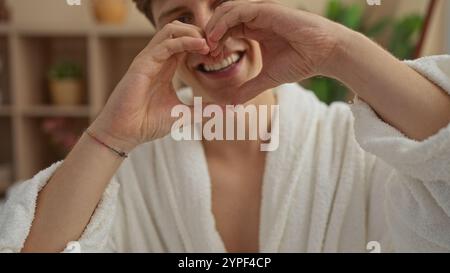 Un giovane in una stanza spa che con le mani crea una forma a cuore, sorride e indossa un accappatoio bianco, che ritrae una sensazione di benessere e relax Foto Stock