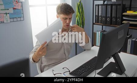 Un giovane si rinfrescava con i documenti in ufficio mentre lavorava al computer, apparendo frustrato e caldo. Foto Stock