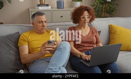 Coppia di mezza età seduta su un divano nel loro salotto, concentrata sui loro dispositivi, con l'uomo che usa uno smartphone e la donna che lavora su un lapto Foto Stock