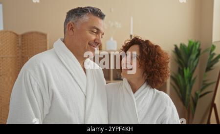 Coppia di mezza età che si diverte insieme in una spa benessere con accappatoi bianchi e sorrisi in un ambiente interno sereno Foto Stock