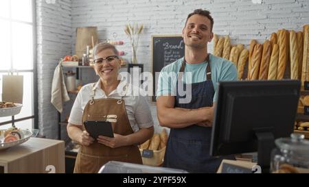 Donna che usa un tablet e uomo con le braccia incrociate sorridente all'interno di un accogliente forno interno con pane sullo sfondo. Foto Stock