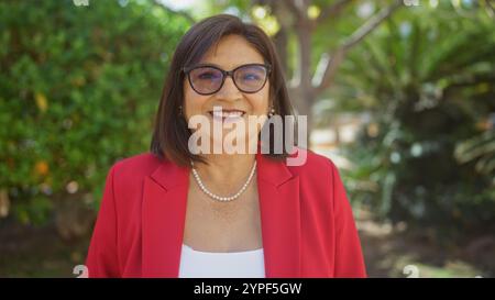 Una bella donna ispanica di mezza età con capelli corti e occhiali sorride calorosamente mentre si trova all'aperto in un parco, vestito con un blazer rosso Foto Stock