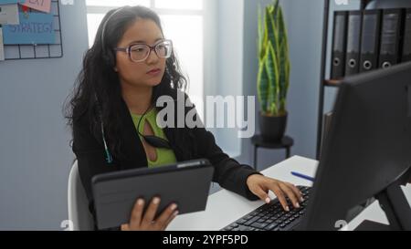Donna che indossa un auricolare che lavora su computer e tablet in un ufficio moderno con file e un impianto in background Foto Stock
