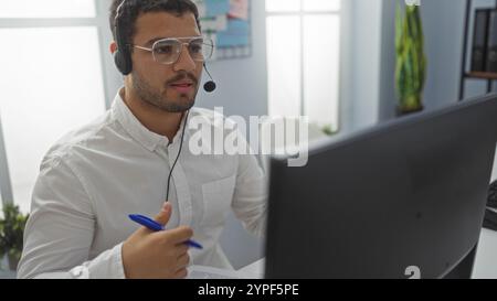 Giovane ispanico che lavora in un ufficio, indossa occhiali e una cuffia durante una chiamata, tiene in mano una penna e si siede alla scrivania con un computer i Foto Stock