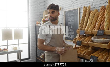 Giovane ispanico con la barba nel negozio di panetteria al coperto che tiene un sacchetto di carta vicino a scaffali di pane e dolci. Foto Stock