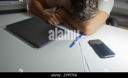 Giovane che riposa la testa su un tavolo con un portatile, un telefono e una penna nelle vicinanze, apparendo stressato in un ambiente domestico rilassato Foto Stock