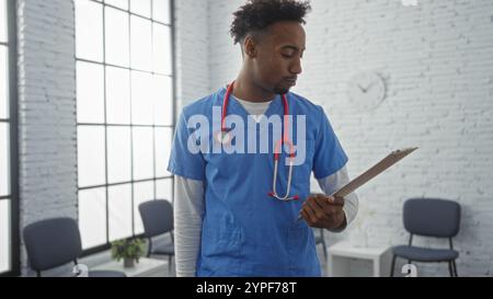 Bel giovane nero professionista di salute maschile con barba in scrub blu che tiene un appunti in una luminosa sala d'attesa clinica. Foto Stock