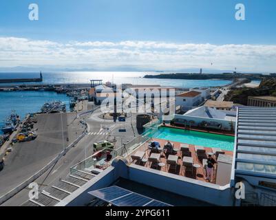 Prospettiva aerea di Tarifa, Spagna, con una terrazza sul tetto con piscina, un vivace porto con barche e un faro lontano sotto un cielo limpido Foto Stock