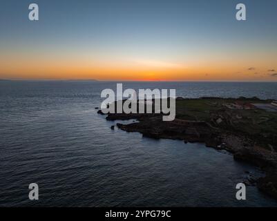 Una vista aerea di Tarifa, Spagna, con un faro su un affioramento roccioso. Il cielo al tramonto mostra sfumature arancioni e blu sulle calme acque marine. Foto Stock