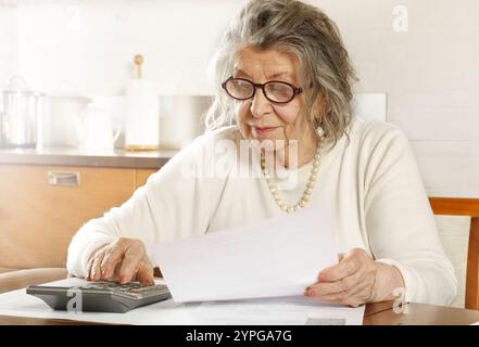 Una donna anziana seduta a un tavolo in cucina che guarda le ricevute con una calcolatrice Foto Stock