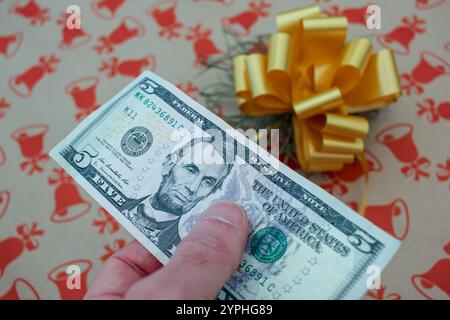 Una banconota da cinque dollari in mano e un regalo di Natale. Concetto, quanti soldi spendiamo per i regali di Natale? Foto Stock