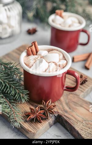 Due tazze in smalto rosso con cioccolata calda, marshmallow e bastoncini di cannella su un tagliere in legno con rami di abete sullo sfondo Foto Stock