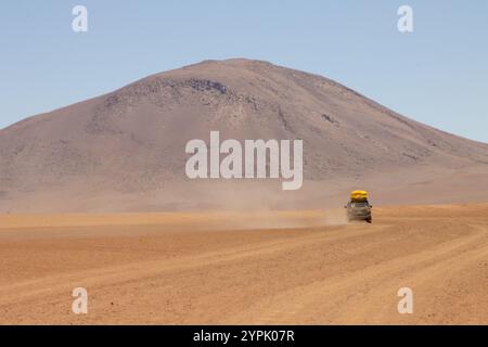 Un veicolo fuoristrada guida nel vasto paesaggio desertico dell'Altiplano boliviano, circondato da aride pianure e maestose catene montuose Foto Stock