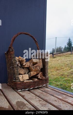 Cestino di vimini con tronchi tritati sul portico della casa. Pila di legna da ardere tagliata. Paesaggio rurale. Tronchi di legno nel contenitore vicino alla cabina. Carrello di tronchi tritati. Foto Stock