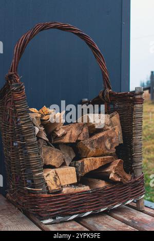 Cestino di vimini con tronchi tritati sul portico della casa. Pila di legna da ardere tagliata. Paesaggio rurale. Tronchi di legno nel contenitore vicino alla cabina. Carrello di tronchi tritati. Foto Stock