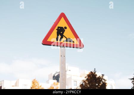 Cartellino triangolare rosso giallo dei lavori stradali su una strada della città contro il cielo blu. Icona di costruzione temporanea in ambiente urbano. Lavori in corso. L'uomo sta lavorando Foto Stock
