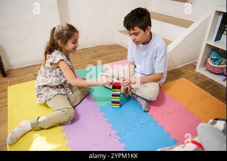 Due bambini seduti su tappeti colorati, impegnati nella costruzione di una torre a blocchi. La scena cattura giocosità e creatività in un ambiente caldo e al chiuso Foto Stock