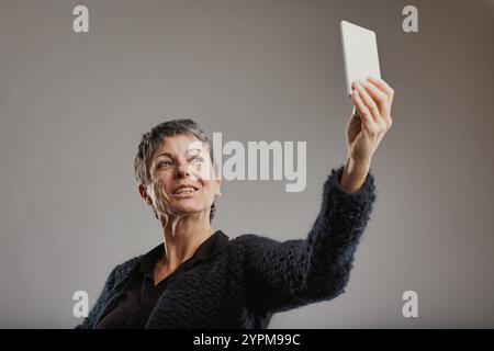 Allegra donna matura dai capelli corti grigi, con un maglione scuro, regge il suo smartphone e scatta un selfie, catturando un momento di gioia e di connessione Foto Stock