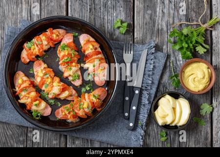 Uunimakkara, cotta in salsicce al forno e condita con formaggio, pomodori e senape in piatto da forno su tavola di legno grigio con ingredienti, classico recip finlandese Foto Stock