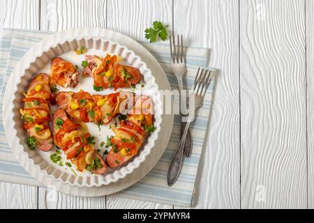 Uunimakkara, al forno con salsicce ricoperte di formaggio, pomodori, senape in piatto da forno su tavola di legno bianco con forchette, ricetta retrò finlandese, orizzonte Foto Stock