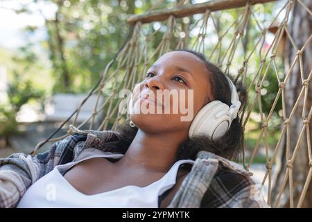 Donna afroamericana che si rilassa nella natura su un'amaca, sceglie la musica su uno smartphone e ascolta le canzoni preferite tramite le cuffie. Foto Stock