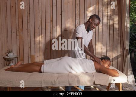 Massaggiatore afroamericano professionista che offre un massaggio rilassante ad una cliente donna, un ospite di lusso del resort glamping. Foto Stock