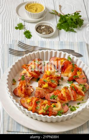 Uunimakkara, cotta in salsicce al forno condite con formaggio, pomodori, coriandolo, senape in piatto da forno su tavola di legno bianco con ingredienti e forchette, pinna Foto Stock