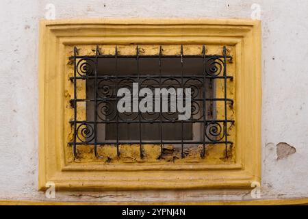 finestra rettangolare adornata da un'intricata griglia in ferro battuto nero, posta all'interno di un telaio in legno giallo intemprato Foto Stock