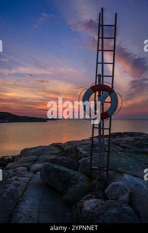 Un tramonto vibrante dipinge il cielo in sfumature di arancione e rosa sopra una tranquilla scena costiera. Un salvagente fissato a una scala metallica si trova su un terreno roccioso Foto Stock