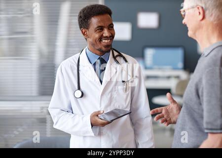 In vita ritratto di un medico afroamericano sorridente che indossa un camice da laboratorio e tiene in mano una compressa mentre parla con un paziente di sesso maschile anziano durante il consulto in clinica, copia spazio Foto Stock