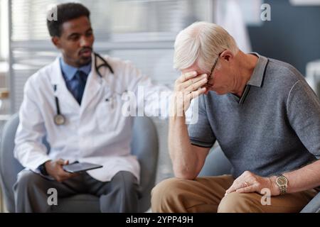 Vista laterale ritratto di un anziano che piange durante la consultazione in clinica dopo aver ricevuto cattive notizie, con il medico in background che lo conforta, copia spazio Foto Stock
