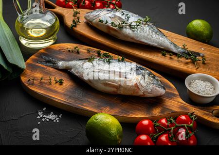 Due pesci dorado freschi e crudi su una tavola di legno su sfondo nero con porri, lime, olio d'oliva e pomodori ciliegini sul tavolo pronti per la cottura. Centro benessere Foto Stock