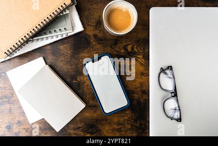 Spazio di lavoro con smartphone in custodia blu, notebook a spirale, carta bianca, bicchieri, tazza da caffè, e un computer portatile su una scrivania in legno - disposizione moderna ed elegante. vista dall'alto Foto Stock