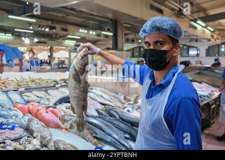Pescivendolo giovane adulto che indossa la maschera, guarda la macchina fotografica, vende pesce fresco bianco hamour sul mercato del pesce tradizionale. Foto Stock