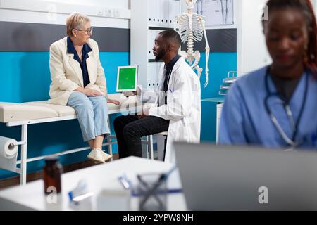 Medico che tiene in mano una compressa con schermo verde spiega i piani di trattamento sanitario al paziente anziano mentre l'infermiera lavora nelle vicinanze. Medico che utilizza un dispositivo digitale con un modello di mockup durante la consultazione con gli anziani. Foto Stock