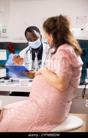 Medico nero che indossa maschera e stetoscopio tiene appunti e consiglia la madre in attesa di mantenere uno stile di vita sano durante la gravidanza. Una donna incinta visita un ostetrico per un controllo di routine. Foto Stock