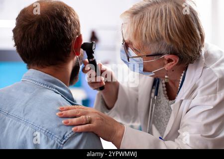 Primo piano del medico anziano che esamina l'orecchio di giovane uomo e discutere i risultati di salute in clinica moderna. Donna anziano medico utilizza da vicino l'otoscopio per controllare l'infezione durante il controllo medico del paziente Foto Stock