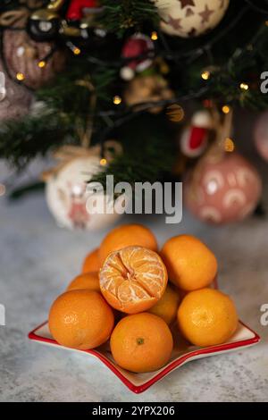 Disposizione festiva di mandarini freschi con un frutto sbucciato in primo piano, posto vicino a un albero di Natale splendidamente decorato Foto Stock