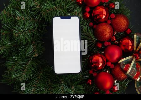 Modello di telefono cellulare con schermo bianco sulla corona di Natale fatta a mano. Festeggiamenti natalizi per il nuovo anno. Decorazioni da cartolina, vista dall'alto, disposizione piatta Foto Stock