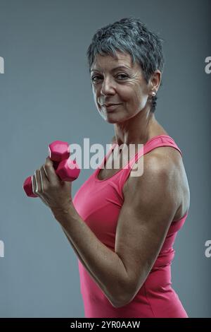 Una donna anziana determinata solleva i manubri rosa, mostrando la sua forza e il suo impegno per uno stile di vita attivo e sano, ispirando gli altri ad abbracciare il fitness Foto Stock