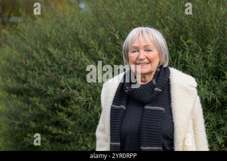 Una donna anziana con i capelli corti grigio-argento e un sorriso delicato sta all'aperto, vestita calorosamente con una sciarpa nera e un morbido cappotto bianco. Foto Stock