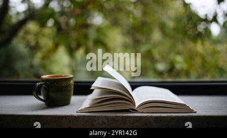 Allestimento Hygge, tazza di caffè e libro aperto vicino alla grande finestra. Concetto di natura morta. Foto Stock