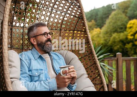L'uomo si sta godendo una tazza di tè caldo, caffè, si sta rilassando a casa, sedendosi su una sedia sospesa sul patio. Foto Stock