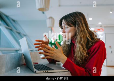 Una donna d'affari frustrata che litiga durante una videochiamata in un ufficio moderno, mostrando un'espressione divertente e usando i gesti delle mani Foto Stock