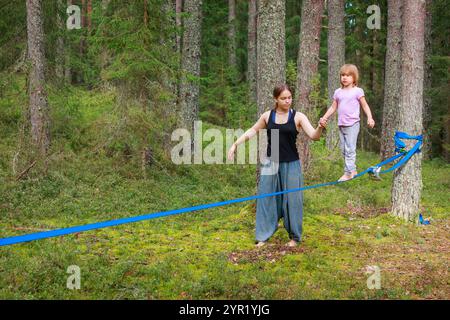 Una ragazza adolescente assiste una giovane cerchia mentre si equilibra con cura su una linea lenta distesa tra due alberi in una foresta tranquilla. La sua postura di supporto Foto Stock