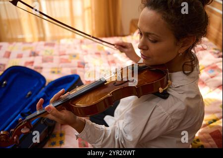 Una donna è profondamente concentrata mentre suona il suo violino, seduta su una trapunta colorata. La luce del sole illumina delicatamente la stanza, creando un bancomat caldo e confortevole Foto Stock