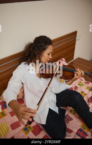 Una donna siede comodamente su un letto mentre suona il violino. L'ambiente e' informale e caldo, riflettendo un'atmosfera rilassata per la pratica della musica o per il perf Foto Stock