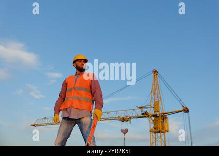 Operaio che tiene in mano una livella a bolla d'aria nel cantiere con gru in background, indossa giubbotto di sicurezza e casco Foto Stock